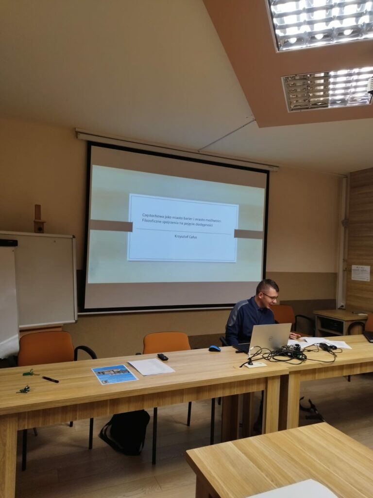 Prelegent siedzący przy biurku na tle prezentacji multimedialnej wyświetlonej na rzutniku.