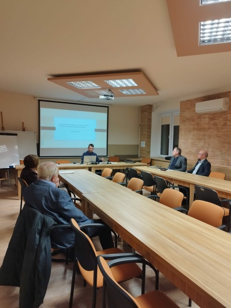 Wszyscy uczestnicy zebrania siedzący przy biurkach ustawionych w podkowę. W tle prezentacja multimedialna wyświetlona na rzutniku.