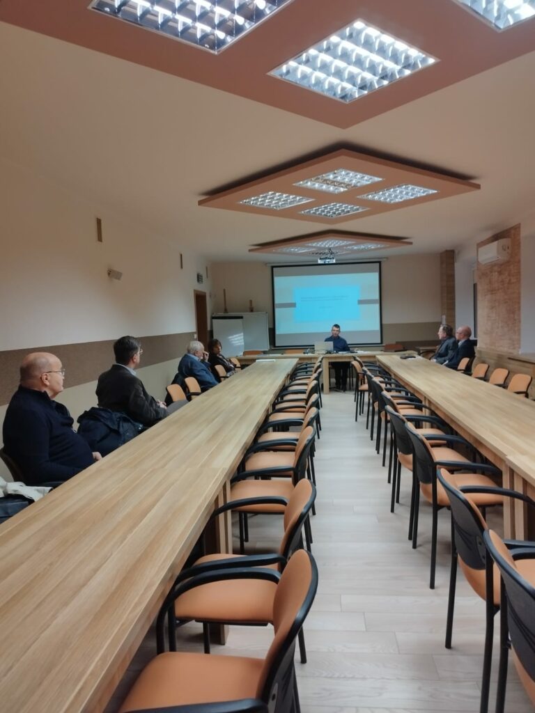 Uczestnicy siedzący przy biurkach ustawionych w podkowę, w tle prezentacja multimedialna wyświetlona na rzutniku. Widok z oddali.