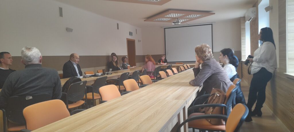Uczestnicy spotkania siedzą przy stołach ustawionych w podkowę. Ja jednym ze stołów stoi osoba trzymająca aparat fotograficzny.
