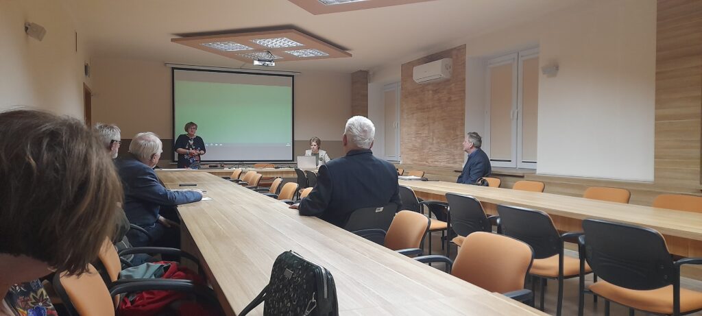 Uczestnicy zebrania siedzą przy stolikach ustawionych w podkowę, zwróceni twarzą do prelegentki i stojącej na środku prezes Julii Dziwoki. Za nią ściana i włączony rzutnik.